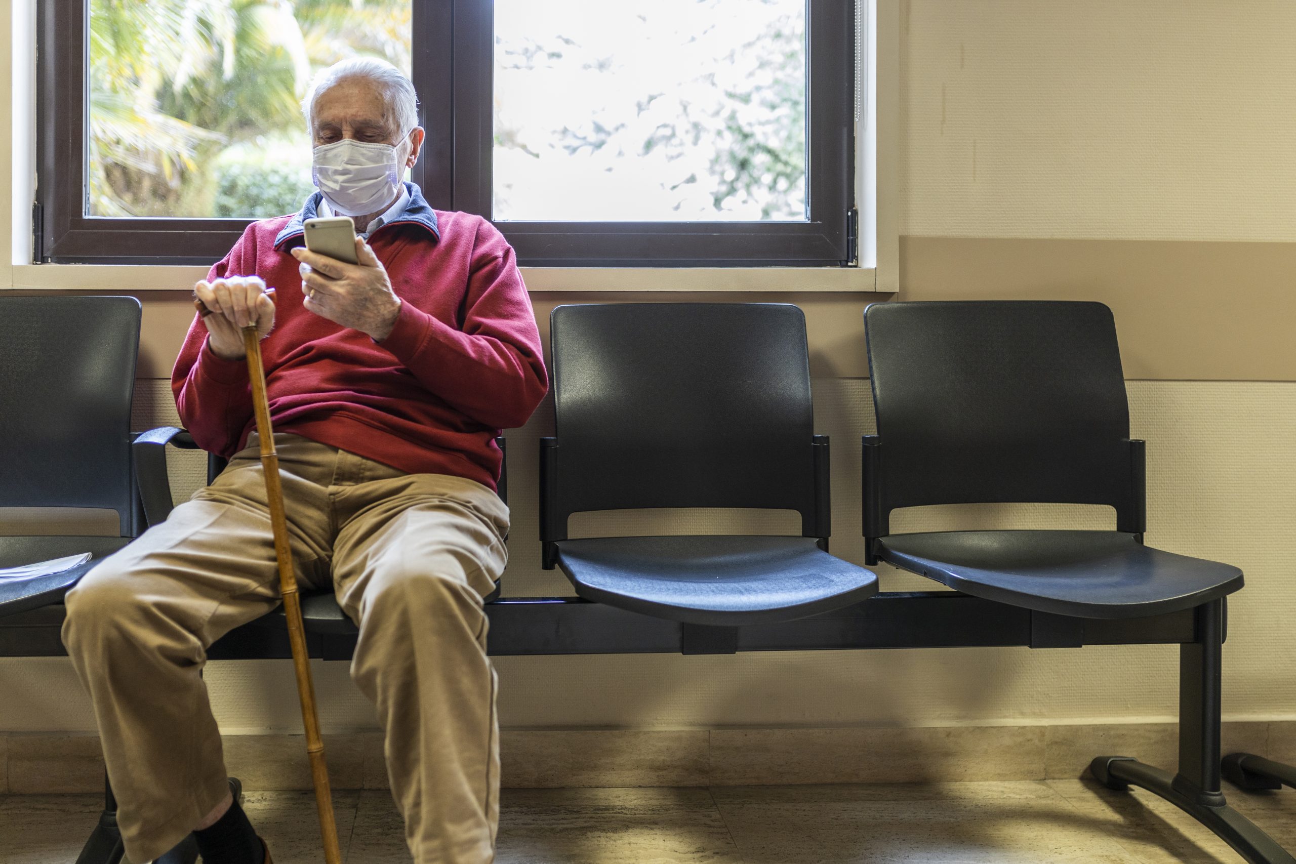 Photo of an elderly male patient in a waiting room using a smartphone.